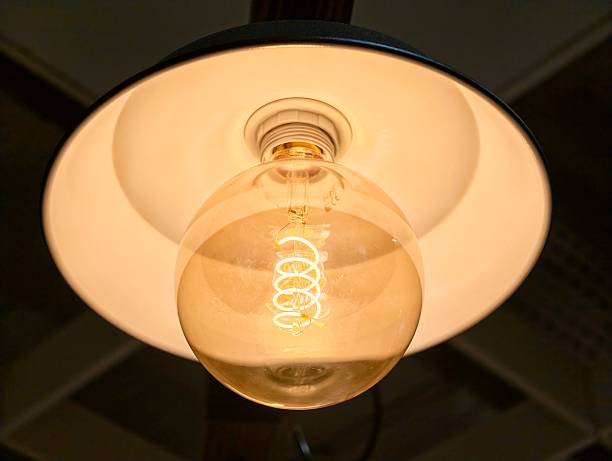 Warm glowing filament bulb under shade, closeup view white metal pendant lamp illuminating textured ceiling