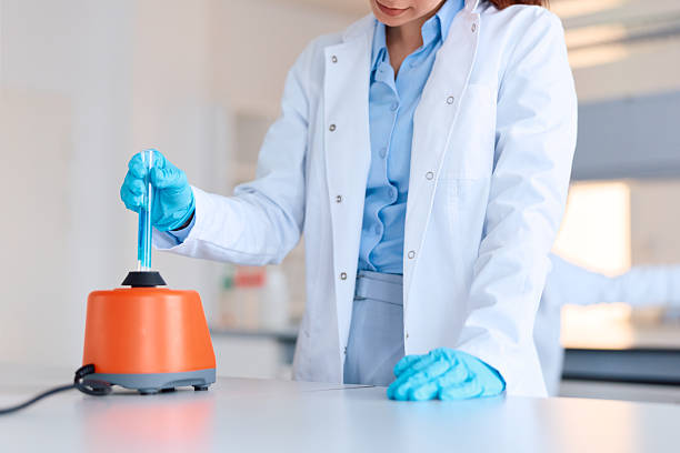 Young adult scientist wearing a lab coat and safety gloves carefully mixing a blue liquid in a test tube using a scientific vortex mixer for various research applications in a modern lab