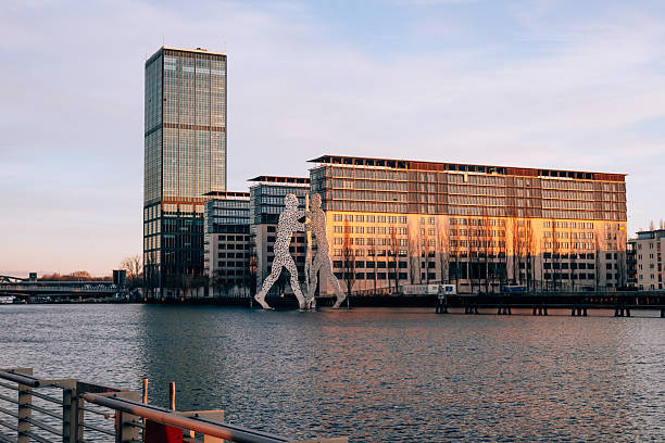 Berlin, Germany – February 15, 2026: The Molecule Man sculpture stands in the River Spree with the Treptowers office complex and surrounding buildings in the background during sunset light. Good weather and clear sky durung the winter.