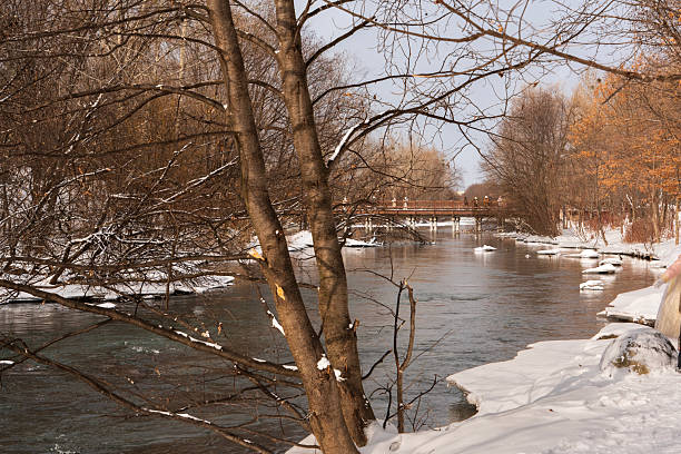 A peaceful winter landscape featuring a flowing river, snow-covered banks, and a wooden bridge among bare trees.