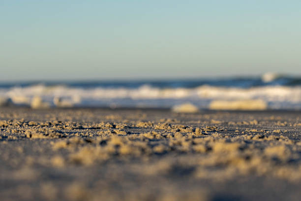 A low-angle, macro-style photograph focusing on the intricate textures of sand clumps on a beach. The foreground is sharply detailed, showing individual grains and small mounds of sand illuminated by the warm, directional light of a setting or rising sun. The background transitions into a soft, creamy bokeh of white surf and blue ocean waves under a clear sky. This tranquil, minimalist composition emphasizes the tactile beauty of the shoreline and is ideal for nature, travel, or mindfulness-themed projects.
