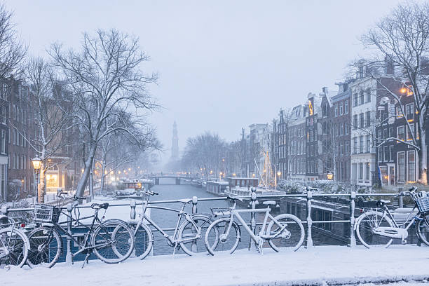 A snow-covered bridge spanning a frozen canal in Amsterdam, with a historic church rising in the background. The winter scene captures a quiet atmosphere and classic Dutch architecture under soft daylight.