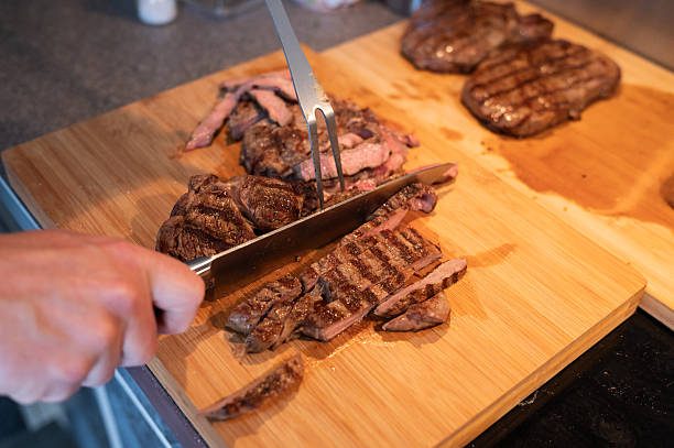 Grilled steak being sliced on wooden cutting board with knife and fork