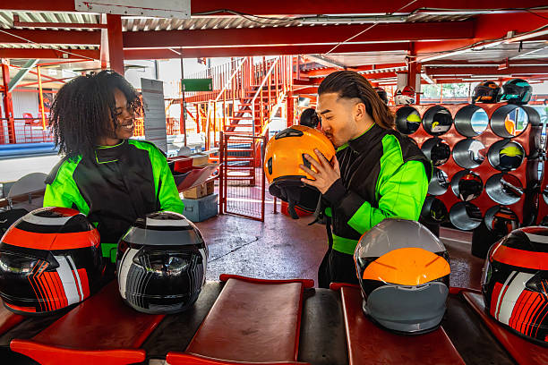 Two young adults in racing suits prepare for a go-kart race at an indoor track, helmets on a table and racks nearby, smiling and bonding before the competition