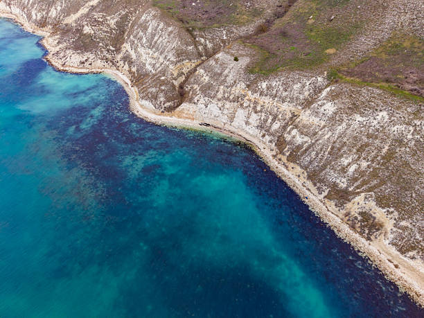 Waves gently hit the rocky shore while clear blue water meets the land at a remote coastline. The land is dry and uneven with some green patches visible.