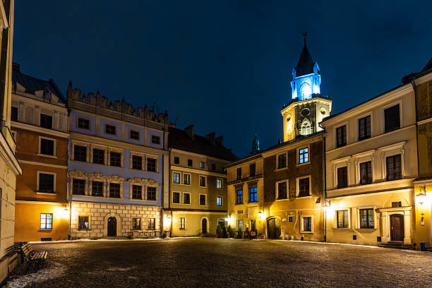 A picturesque night view of the Market Square (Rynek) in Lublin's Old Town. The scene features the iconic Konopnica Tenement House (Kamienica Konopniców) with its ornate blue facade and the towering Trinitarian Tower (Wiea Trynitarska) illuminated with a striking blue light in the background. The empty cobblestone square and warm ambient lighting of the historic buildings capture the serene and mystical atmosphere of this Polish heritage site.