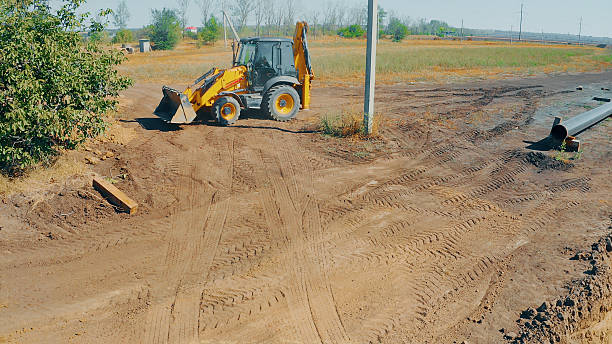 Yellow backhoe loader parked on a dirt construction site with tire tracks and pipes