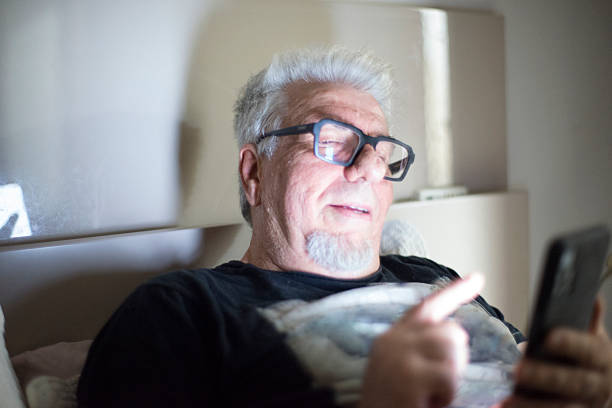 A 70-year-old man lying comfortably in his bed at night, interacting with a smartphone. The scene portrays connectivity in old age, whether for leisure, communicating with family, or accessing information, highlighting technology as part of the daily lives of the elderly.