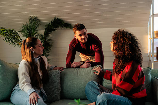 Three young friends gathered around a sofa in a cozy living room, engaged in friendly conversation. One man leans casually over the back of the couch while the two women sit facing him, smiling and talking.