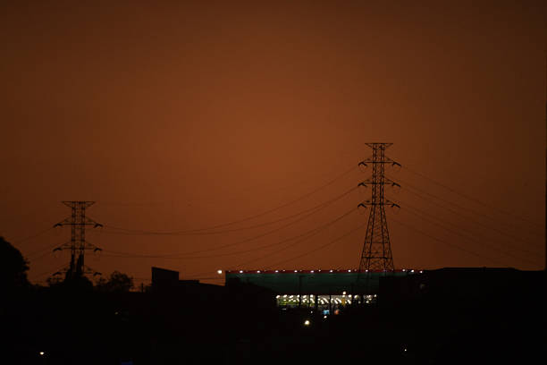 Night photograph with a reddish tone showing a large illuminated smokestack and two nearby transmission towers, representing industry, energy production, and urban infrastructure.