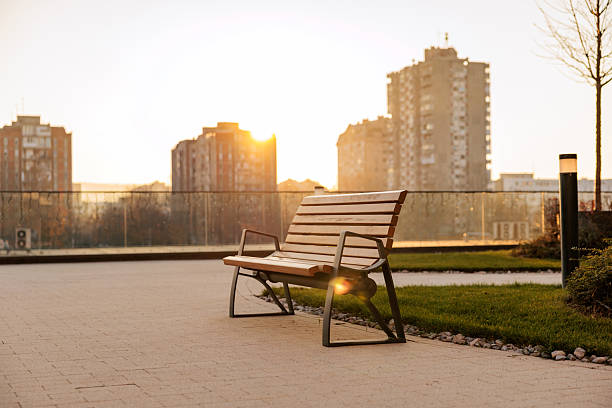 Wooden bench standing on a paved walkway in a modern residential complex, illuminated by warm sunset light. City apartment buildings rise in the background as the sun sets behind them, creating a calm and atmospheric scene.