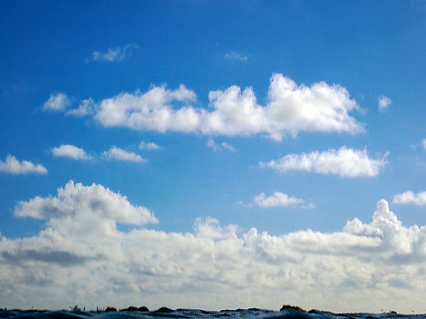 A serene landscape image features rolling sandy dunes covered in patches of green grass and low vegetation along a coastal beachfront. The foreground shows textured sand and natural dune formations, transitioning to the calm ocean horizon. Above, a clear blue sky dominates with fluffy white cumulus clouds drifting peacefully. No people are present, emphasizing the untouched natural beauty of the seaside environment. This image captures a tranquil daytime scene ideal for commercial uses such as travel brochures, environmental campaigns, wellness advertisements, or backgrounds in media content promoting relaxation and nature escapes. The composition highlights the expansive sky and earthy tones of the dunes, suitable for editorial or promotional materials in tourism, real estate, or outdoor lifestyle sectors.