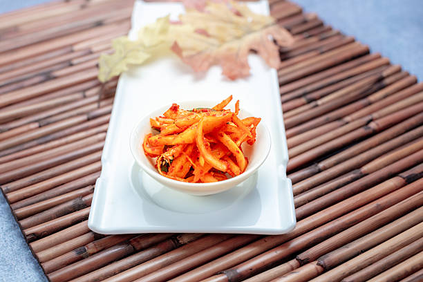 The Dish: Doraji Muchim (Seasoned Bellflower Root)This classic Korean side dish is made from the roots of the Platycodon (balloon flower), known for its unique bittersweet flavor and crunchy, fibrous texture. It is a favorite for adding a refreshing zing to any meal.The Flavor Profile: The natural bitterness of the bellflower root is carefully balanced with a vibrant dressing made of gochugaru (red chili flakes), vinegar, sugar, and garlic, creating a perfect harmony of spicy, sweet, and tangy notes.Health Benefits: In Korea, bellflower root has long been used in traditional medicine to support respiratory health and boost the immune system.The PresentationVibrant Colors: The bright orange-red seasoning makes the dish visually striking, especially when served in a clean white ceramic bowl as shown in the photo.Rustic Elegance: Set on a white long platter over a traditional bamboo mat (Bal) and decorated with autumn leaves, the presentation reflects a sophisticated, seasonal aesthetic.Appetite Stimulator: Its sharp, tangy taste makes it an excellent \