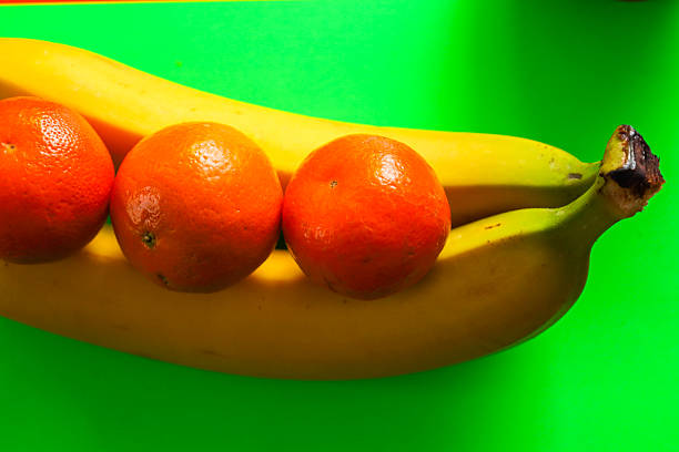 Three ripe orange mandarins lie on two yellow bananas, creating a bright fruit composition on a rich green background. This tropical fruit still life symbolizes healthy eating, freshness, and a vitamin boost.