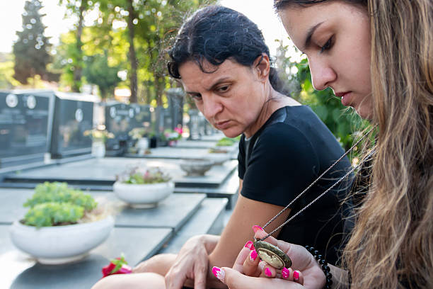 Mother and young adult daughter sitting at a cemetery grave, finding solace in a locket, grieving the loss of a family member and supporting each other during a difficult time
