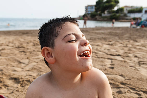 Young boy with closed eyes laughing joyfully on sand, experiencing happiness and summer fun at seaside