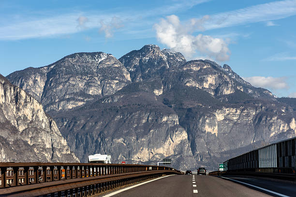 Modern highway road leading through impressive rocky mountain landscape under blue sky. Scenic alpine view with vehicles driving toward dramatic peaks. Concept of travel, transportation, freedom and journey across Europe.