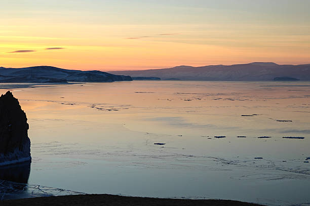 Golden-pink sunset washes over a tranquil frozen lake with scattered ice floes, a dark rocky cliff in the foreground and soft mountain silhouettes on the horizon, evoking calm and solitude.