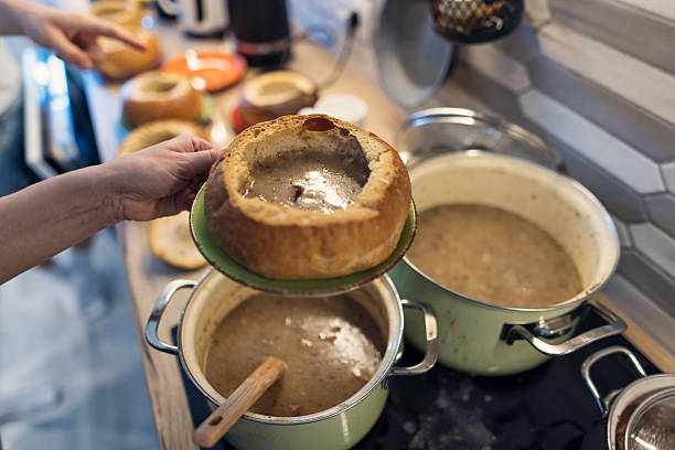 Preparation for traditional Easter breakfast.Mother pours hot Easter rye soup (żurek) into a hollowed bread bowl held on a green plate and hands it to the son. The thick, hearty soup is served straight from the pot in a cozy kitchen setting, with utensils and hanging baskets in the background adding to the warm, home‑cooked atmosphere.Shot with Canon R5