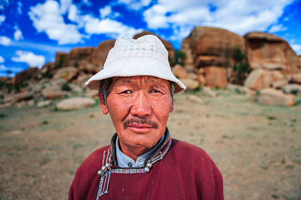 A close-up portrait of an older man standing outdoors in front of rounded granite boulders and a bright blue sky with scattered clouds. He wears a white knit hat and a maroon traditional robe with decorative fastenings; his weathered face, short mustache and thoughtful expression are sharply in focus while the rocky landscape behind him is softly blurred.