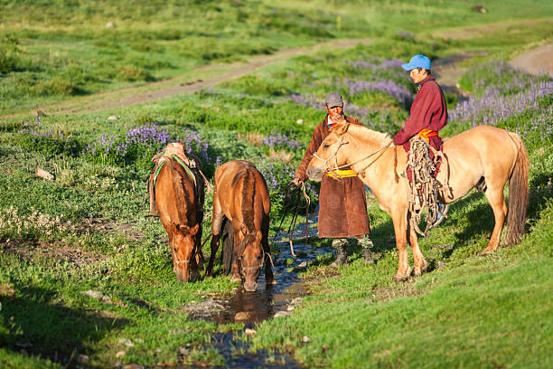 Two riders with horses pause beside a narrow stream in a sunlit green valley. Two chestnut horses drink from the water while a rider on a pale dun horse holds their reins; another person stands nearby in a long brown coat and cap, smoking. Purple wildflowers and rolling grassy slopes fill the background, creating a calm, pastoral scene.