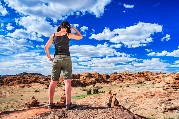 A person stands on a rocky outcrop with their back to the camera, hand raised to shield their eyes while surveying an expansive field of orange-red sandstone formations and scattered shrubs. Small stacked stone cairns sit in the foreground on sun-warmed rock. A vivid deep-blue sky dotted with bright white clouds stretches overhead, emphasizing the wide-open, rugged landscape and a feeling of adventure.