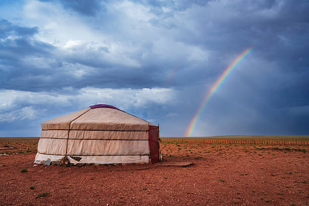 A solitary traditional yurt sits on a wide red-earth plain beneath heavy, brooding clouds. A vivid rainbow arcs across the sky to the right, with a faint secondary band visible, while low vegetation and a distant fence stretch to the horizon, conveying the vast, open steppe and a moment of shifting weather.