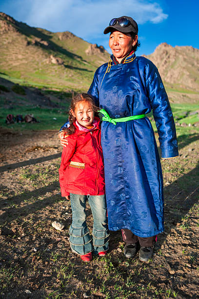 A smiling woman in a deep blue traditional robe with a bright green sash stands with her arm around a young girl wearing a red jacket and ruffled denim pants. They are bathed in warm sunlight on rocky, grassy ground with sunlit hills and a clear blue sky behind them, conveying a candid moment of family and rural life.
