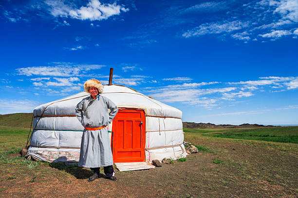 A person wearing a long traditional robe (deel) tied with a bright sash and a large fur hat stands with hands on hips in front of a white yurt with a vivid orange door. The yurt sits on open grassy steppe with low rolling hills beneath a deep blue sky scattered with thin clouds, evoking a quiet, rural nomadic scene.