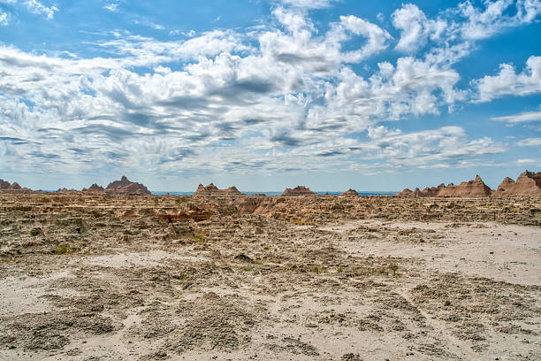 A desert landscape with a few small hills and a large body of water in the distance. The sky is mostly clear with a few clouds scattered throughout