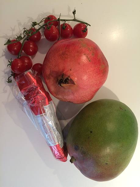 A colourful arrangement of fresh mango, pomegranate and tomatoes alongside a wrapped bar of chocolate, displayed on a kitchen counter. The combination of fresh fruit and confectionery creates an interesting and appealing food still life composition.