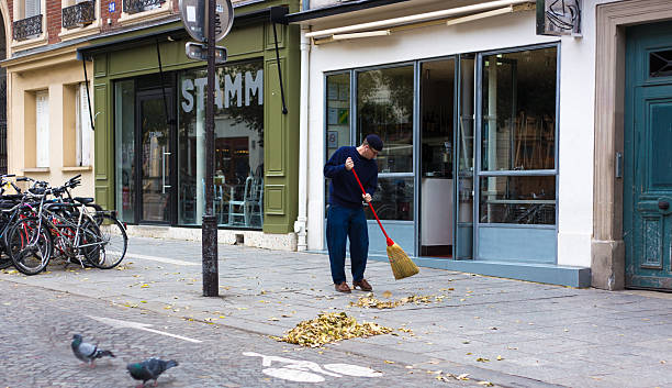Paris, France - October 29, 2025: A man sweeps leaves on a calm sidewalk (Rue Jean-Pierre Timbaud) in the 11th arrondissement early in the morning.