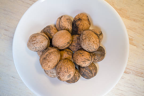 A collection of uncracked walnuts sits in a white bowl placed on a wooden table. The nuts have a natural brown shell and vary in color and texture.