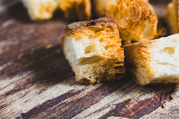 Small pieces of bread are set on a wooden surface. The pieces show uneven cuts and color changes. This scene is likely part of a cooking or baking process in a kitchen.