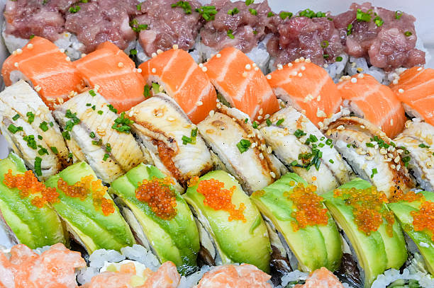 Assorted sushi set displayed in rows, featuring various toppings. Includes salmon (Salmo salar) with orange hue, tuna (Thunnus spp.) with a red/purple appearance, eel (Anguilla spp.) with a grilled surface, and avocado (Persea americana) presenting a green, smooth texture. Decorated with orange caviar, offering color contrast. Garnished with chopped green onions and sesame seeds, enhancing flavor and visual appeal. Each sushi piece set on a base of vinegared rice, arranged on a light-colored tray, highlighting the vibrant ingredients.