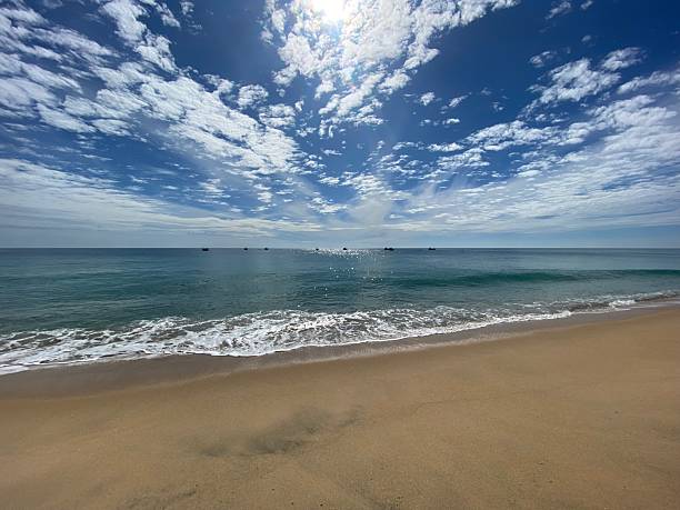 Wide-angle view of a peaceful sandy beach with gentle waves rolling onto shore under a vibrant blue sky filled with scattered white clouds. Sunlight reflects on calm ocean water, while small boats rest on the horizon. Ideal for concepts of summer travel, relaxation, tropical vacation, coastal tourism, and natural beauty.