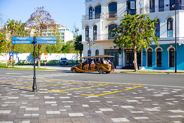 A group of tourists rides in a vintage yellow open-top electric car along a cobblestone street surrounded by modern white buildings and trees. Phu Quoc, Vietnam, December 28, 2025