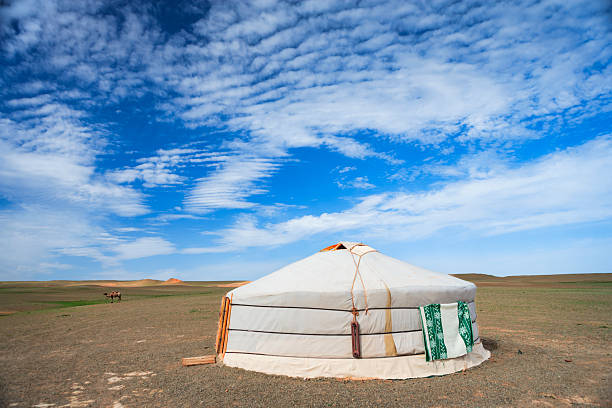 A traditional white yurt sits alone on a wide, flat plain beneath a vast blue sky streaked with textured, rippling clouds. A lone camel grazes in the distance while low dunes and sparse vegetation mark the horizon; a green patterned cloth hangs on the yurt’s exterior.