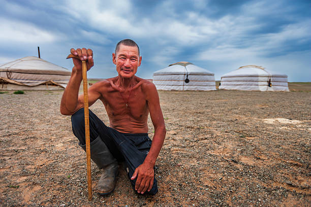 A shirtless older man crouches on gravelly ground, holding a carved wooden staff and smiling gently at the camera. He wears dark trousers tucked into sturdy boots and a small pendant around his neck. In the background three white felt gers (yurts) sit on the open, arid plain beneath a wide, dramatic cloudy sky, conveying a remote pastoral campsite on the steppe.