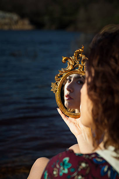 Conceptual outdoor portrait of a young woman holding an ornate vintage mirror that reflects her face against a natural water background. The composition highlights themes of identity, self-reflection, introspection, and personal awareness. Warm natural light enhances the golden frame details while the soft background creates depth and focus on the reflection. Ideal for psychology concepts, self-discovery themes, personal growth content, lifestyle branding, editorial storytelling, and creative advertising projects.