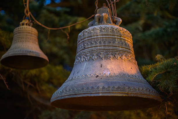 Two ornate metal bells hanging outdoors on ropes among green tree branches at dusk, warm natural light, shallow depth of field, rustic religious atmosphere.