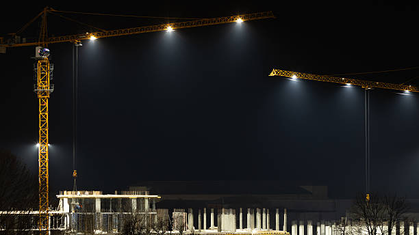 Night view of a construction site with two tall cranes illuminating a concrete structure. Light fog, cold atmosphere, urban industrial scene, and winter night concept.