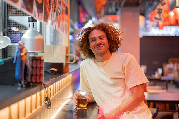Smiling young man with curly hair enjoying a casual drink at a lively izakaya bar, relaxed and content in warm evening light, reflecting urban nightlife and social leisure
