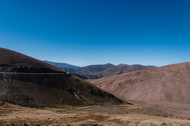 A vast, rocky desert landscape with a clear blue sky above. The mountains in the distance create a sense of depth and scale, while the barren terrain suggests a harsh and unforgiving environment