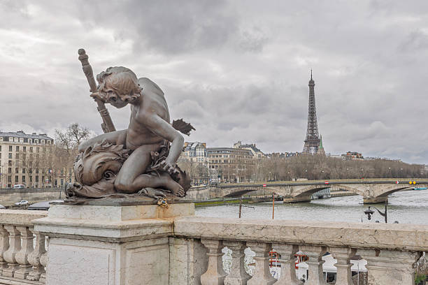 Bronze cherub sculpture on the ornate Pont Alexandre III overlooking the Seine River in Paris, France. The Eiffel Tower rises in the background beyond a stone bridge and classic Haussmann buildings under a dramatic cloudy sky. Captured in winter light, this image highlights iconic Parisian landmarks, Belle Époque architecture, and European travel destinations. Ideal for themes of romance, tourism, French culture, historic bridges, and cityscape photography.