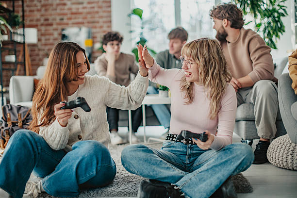 Young women sitting cross legged on floor holding controllers and smiling at camera in natural daylight.