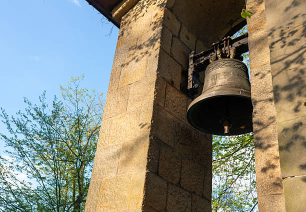 Close view of a bronze bell fixed to an iron yoke within a stone tower frame, lit by warm sunlight. Concept image for craftsmanship, old architecture detail, religion, and traditional city heritage.