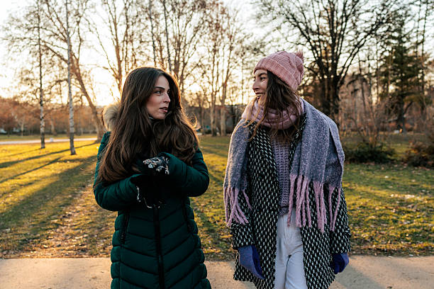 Two young women dressed in warm winter coats, scarves, and gloves walking side by side along a park path during autumn. They are engaged in conversation, looking at each other while enjoying the soft golden sunset light.