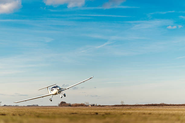 White single-engine light aircraft with long wings lifting off a grassy airfield into a clear blue sky on a sunny day, wheels retracting as it begins its flight