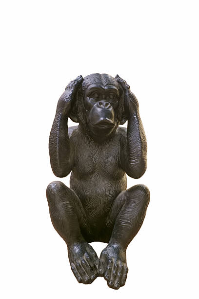 A sculpture of a monkey sits on a plain background. The figure has hands covering its ears and a thoughtful expression. It is made of dark material and shows detailed features.
